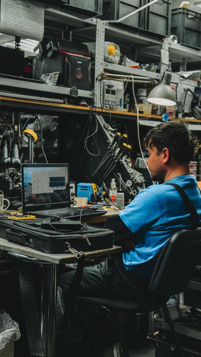 Engineer focused on laptop in cluttered workshop. Ideal for tech and industry themes.